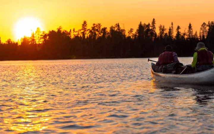 two people sit in a canoe on calm water while the sun sets behind trees on the shore